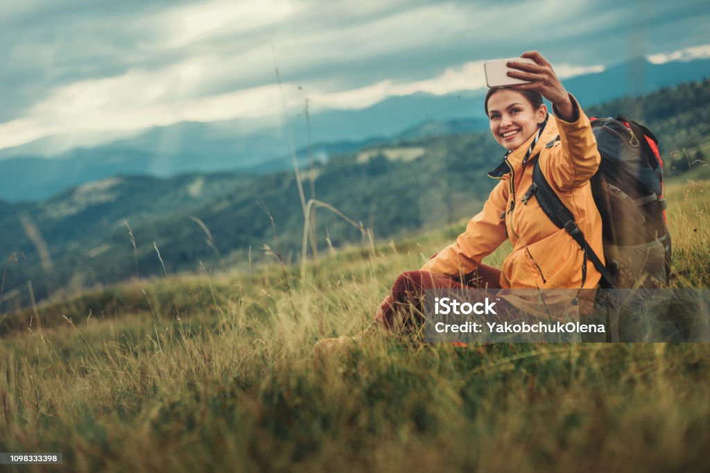 Joyful nice woman making selfies from the mountain hill Positive attractive young woman sitting on the mountain hill while making panorama photos Hiking Stock Photo