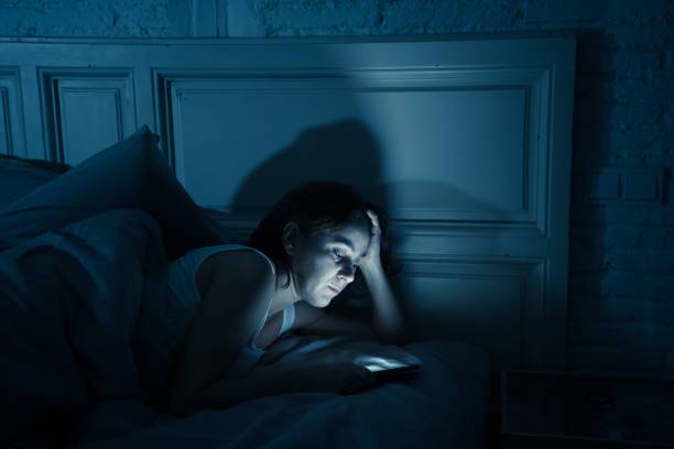 joven atractivo despierta tarde en la noche usando el teléfono inteligente en la cama en un dormitorio oscuro. usando el móvil para chatear y enviar mensajes en adicción a internet, móvil abuso y concepto de insomnio - working bed smart phone bedroom fotografías e imágenes de stock