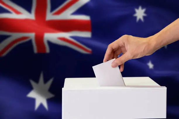 Photo of Close-up of human hand casting and inserting a vote and choosing and making a decision what he wants in polling box with Australia flag blended in background.