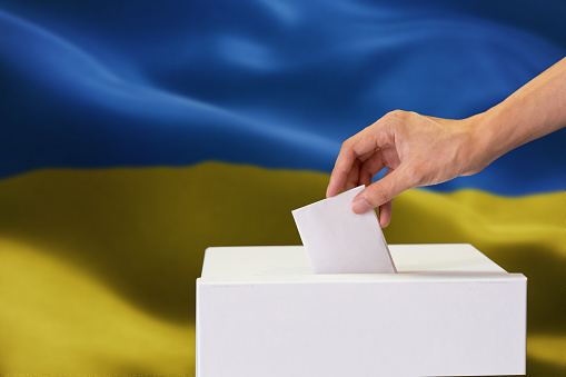 Close-up of man casting and inserting a vote and choosing and making a decision what he wants in polling box with Ukraine flag blended in background.