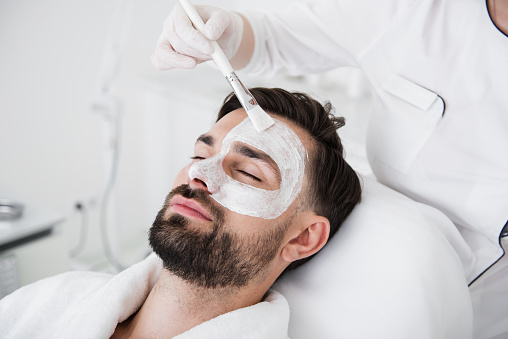 Relaxed bearded young man being in spa salon and professional cosmetologist putting white clay mask on his face with a brush