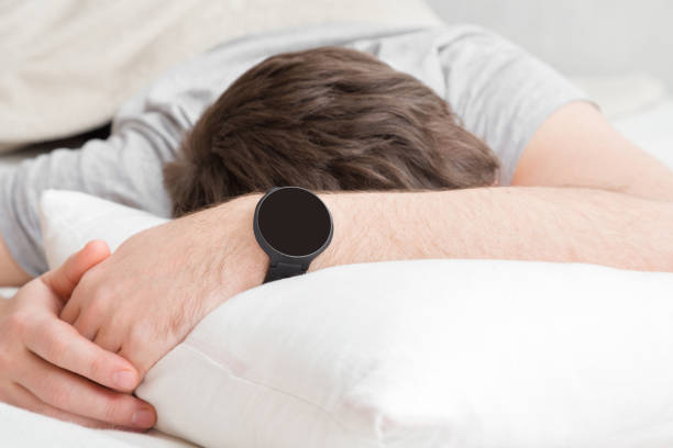 istock Young man's head lying on white pillow in bed. Modern, black smart watch on wrist. Alarm clock. Not getting enough sleep. Can't wake up. Place for digital numbers, logo or mobile app on empty screen. 1098351782