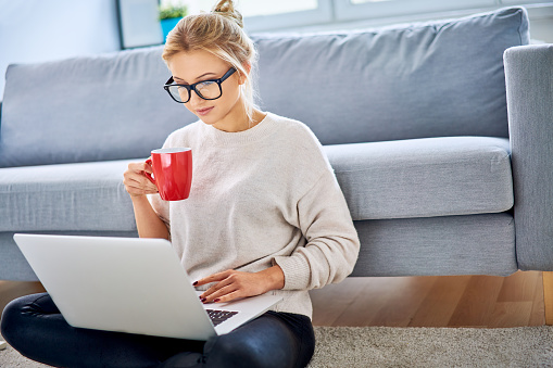 Young woman having coffee while using laptop and sitting on floor at home