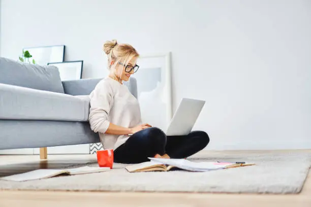Photo of Female student sitting on floor of her apartment with laptop and notes studying
