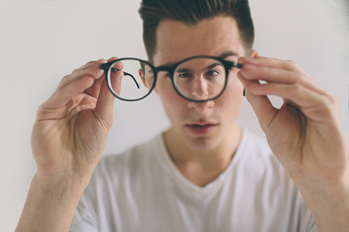 Closeup portrait of young man with glasses. He has eyesight problems and is squinting his eyes a little bit. Handsome guy is holding his eyeglasses right in front of camera with one hand. The concept is isolated on a white background
