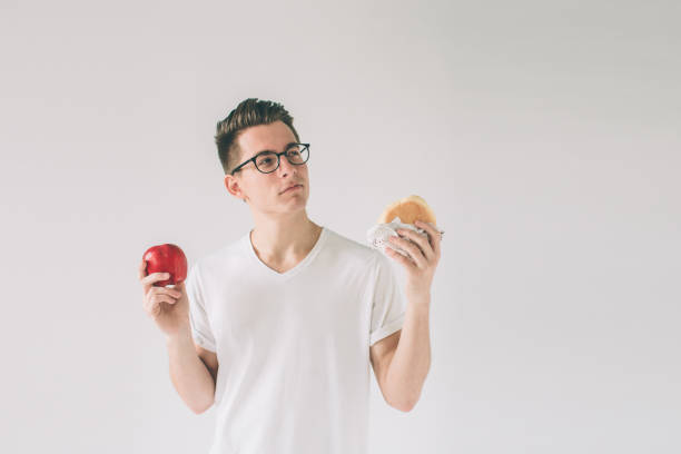 istock Bearded man in a white shirt on a light background holding a hamburger and an apple Man makes the choice between fast and healthy food. Tasty or useful The dilemma choosing a different lifestyle. Nerd is wearing glasses 1098366616