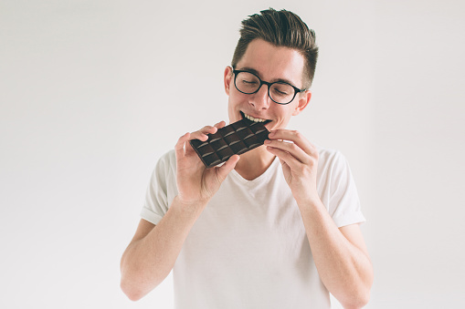 young man eating a chocolate bar. Nerd is wearing glasses