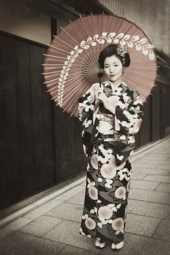A stock photo of a beautiful young girl dressed in traditional Japanese Kimono dress and parasol while standing on a quiet street in Kyoto, Japan.