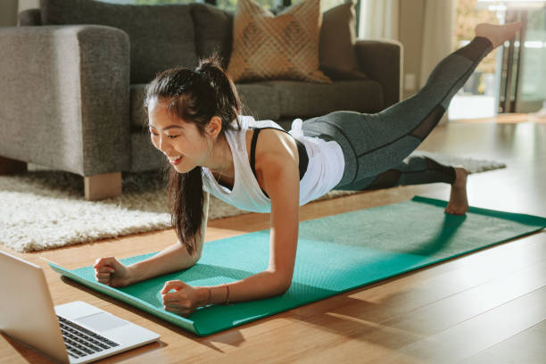 Woman watching sports training online on laptop. Smiling woman exercising at home and watching training videos on laptop. Chinese female doing planks with a leg outstretched and looking at laptop. exercise mat photos stock pictures, royalty-free photos & images