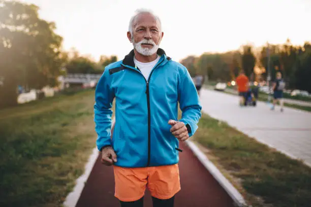 Senior bearded man jogging on a running track outdoors, in a tracksuit.