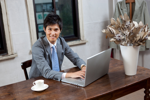 A stock photo of a handsome young businessman smiling while working on a laptop in an office space.