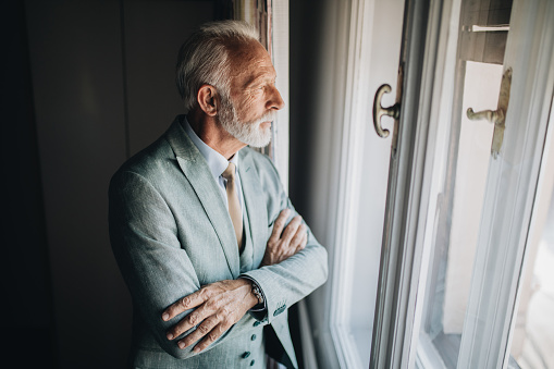 Fashionable senior businessman in suit, standing in his hotel apartment by a window.