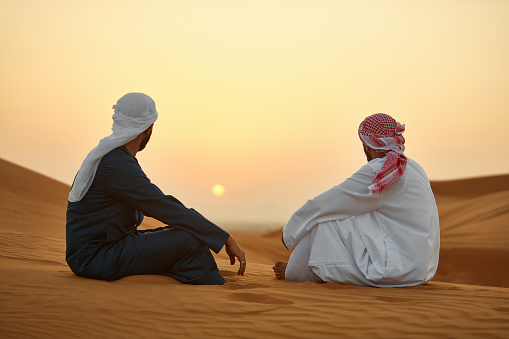 Male friends looking at sunset while sitting on sand. Mid adult men are traveling in desert. They are in traditional wear.