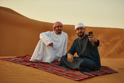 Man taking selfie with friend on smart phone. Arab males in traditional wear are sitting on carpet. They are at desert.