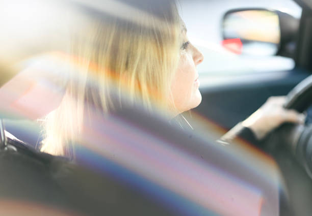 Attractive woman in her 60’s sits behind the wheel of a vehicle on a sunny day with her eyes on the road and her hands at the wheel. Candid image of a cheerful woman driving