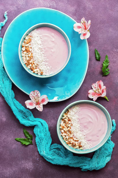 Healthy Breakfast-yogurt with almonds and coconut flakes in a blue bowl on a purple background decorated with flowers of Alstroemeria. View from above