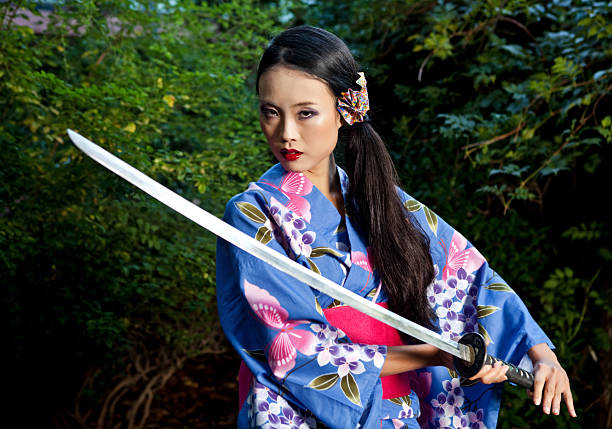A woman with long hair stands in the woods while holding a samurai sword.  She is posing as a geisha entertainer wearing traditional Japanese attire with the katana.