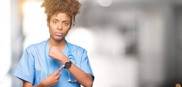 молодой афро-американский врач женщина за изолир�ованным фоном в спешке указывая, чтобы посмотреть время, нетерпение, расстроен и зол на сро - checking the time women impatient wristwatch стоковые фото и изображения