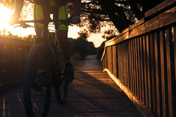 backlit cyclist on a mountain bike runs on a wooden bridge of a rural road, the sun illuminates the rider. the golden light illuminates the wood and leaves the trees in silhouette. Dark mood
