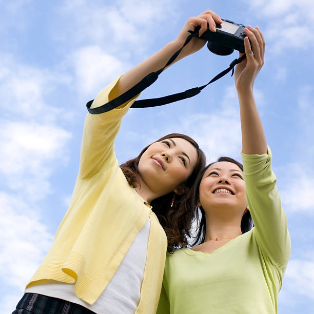 Two young Asian women smile while photographing themselves with a camera outdoors.