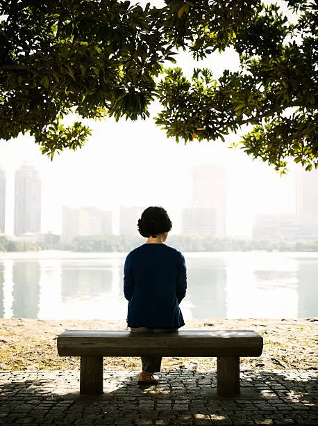 Rear view of Asian senior woman sitting on a park bench under the shade of two large trees looking at view of lake and buildings on other side.   