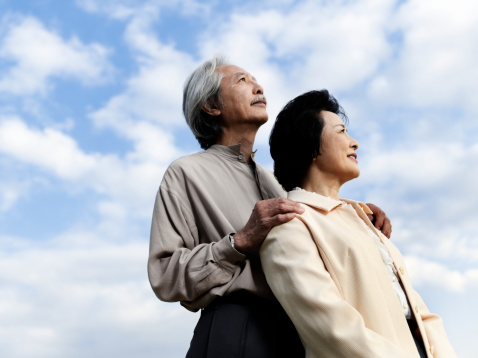 Senior Asian couple hold hands while taking a stroll along sandy beach at waters edge on a cool day.   Male has grey hair wearing eyeglasses and jacket and scarf.  He is turned smiling at wife as they walk.   Female wears jacket and skirt.  The couple continue bonding and enjoying their retirement lifestyle.  Blue sky and hazy horizon.  Horizontal shot.