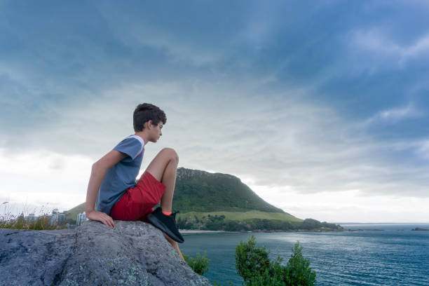 Boy sits on rock on Leisure Island looking out to sea with Mount Maunganui  under evening light and dark clouds in background