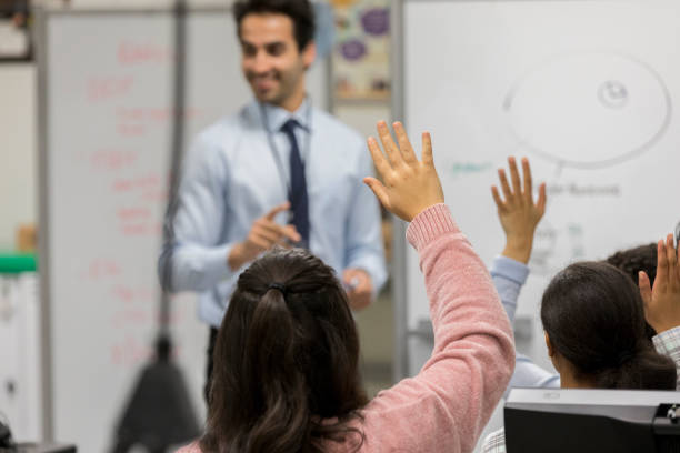 Student raises hand during class Rear view of unrecognizable teenage girl raising her hand to ask or answer a question during science class. A male teacher is in the background. teenage high school girl raising hand during class stock pictures, royalty-free photos & images