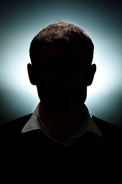 A shadowy silhouette of a man in a sweater vest A portrait of a man lit with only a hair light and a background light. obscured face photos stock pictures, royalty-free photos & images