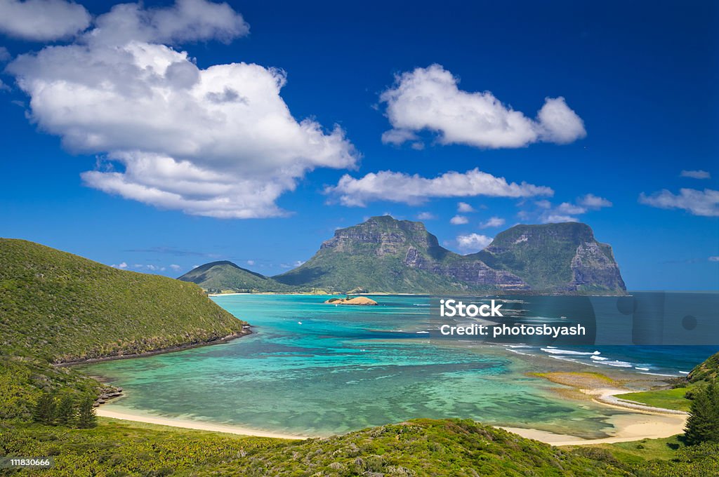 View over Lord Howe Island lagoon to Mt Gower, Australia View south over the stunning turquoise lagoon to the peaks of Mts Lidgbird and Gower. Taken from Mt Eliza on Lord Howe Island, Australia. Fluffy clouds stand out from the deep blue sky overhead. This tiny island is a delightful tourist destination. Lord Howe Island Stock Photo