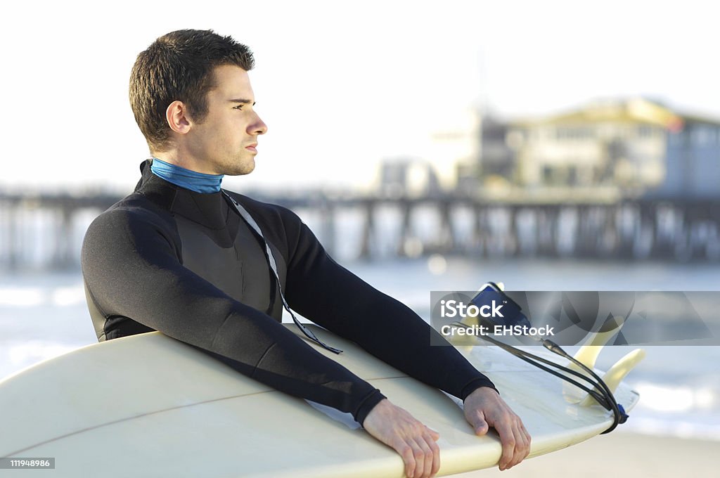 Giovane uomo con tavola da surf nella muta da sub e molo di sfondo - Foto stock royalty-free di Acqua