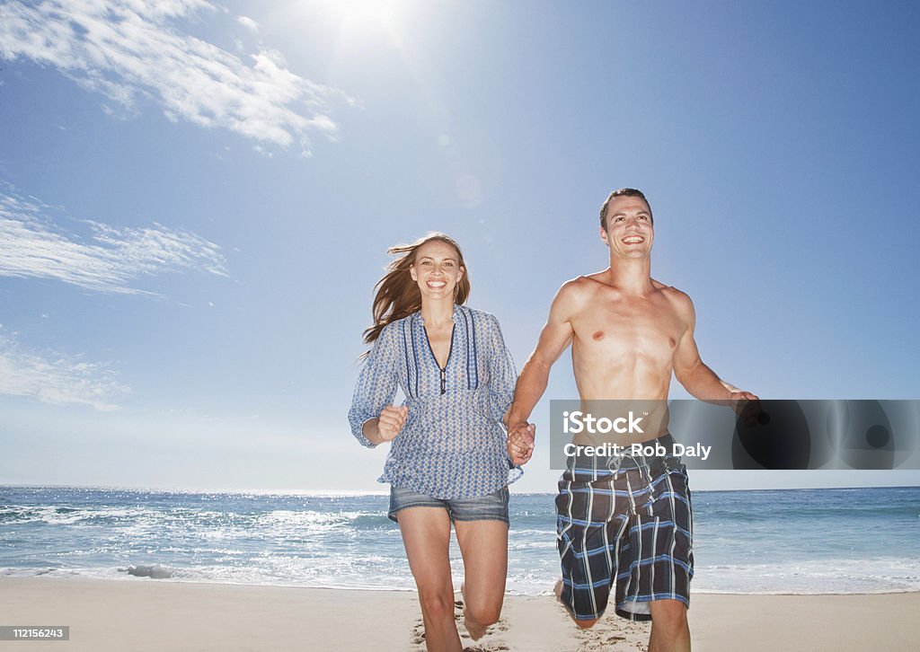 Coppia in esecuzione sulla spiaggia sorridente e tenendo le mani - Foto stock royalty-free di 20-24 anni