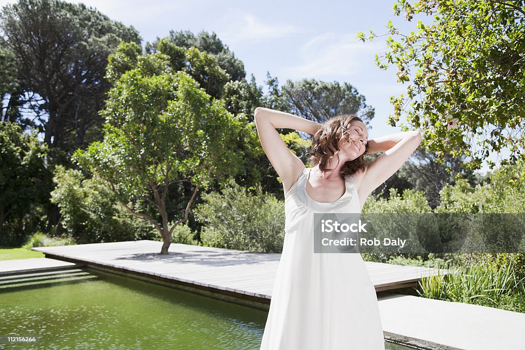 Mujer sonriente de pie cerca de la piscina con las manos detrás de la cabeza - Foto de stock de 30-39 años libre de derechos