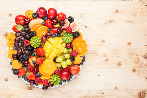 Healthy fruit platter, strawberries raspberries oranges plums apples kiwis grapes blueberries mango persimmon on wooden table, top view, copy space for text, selective focus