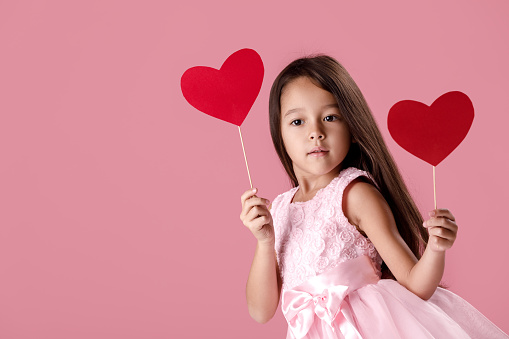 portrait of a cute little girl in a pink dress holding two paper heart on a pink background. St. Valentine's Day