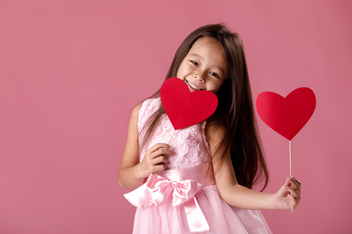 portrait of happy cute little girl in a pink dress holding two paper heart on pink background. St. Valentine's Day