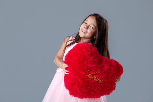 portrait of smiling cute little girl in pink dress holding big red heart on gray background. St. Valentine's Day