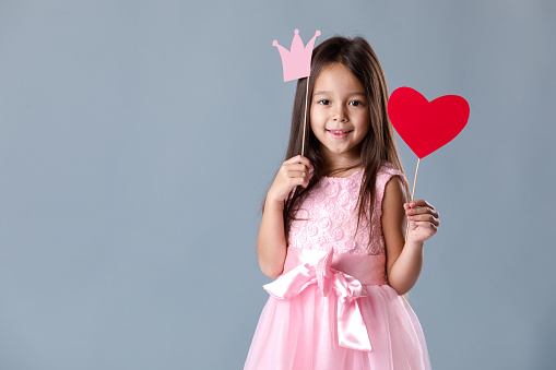 portrait of a cute little girl in pink dress holding paper heart and princess crown on gray background. St. Valentine's Day