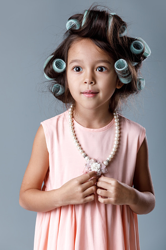Funny cute little child girl in pink dress and hair curlers posing on gray background. Human emotions and facial expression