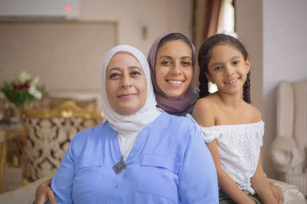 A mother, daughter and grandmother are sitting together in their living room. They are smiling at the camera.