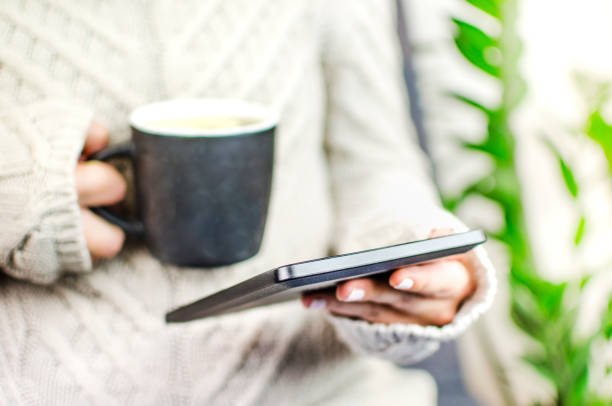 Female person wearing sweater holding electronic reader and a hot beverage, enjoying leisure time at home