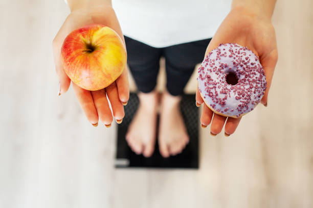 régime alimentaire. femme de mesure corps poids sur pesant échelle holding donut et apple. sucreries sont malsaines de la malbouffe. restauration rapide - food measuring hamburger dieting photos et images de collection