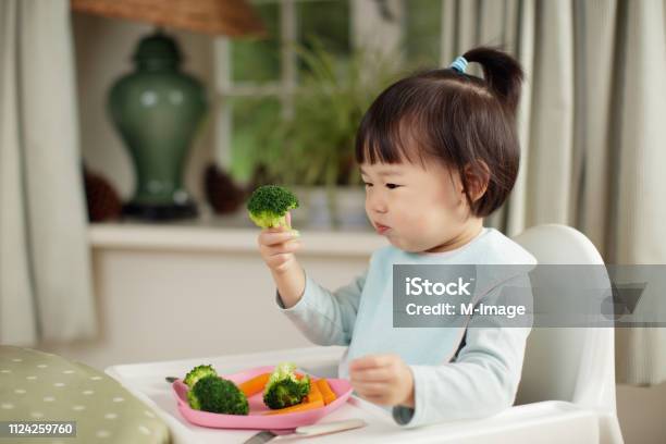 Kleinkind Mädchen Essen Gesundes Gemüse Sitzen Auf Hohen Stuhl Neben Einem Esstisch Zu Hause Stockfoto und mehr Bilder von Essen - Mund benutzen