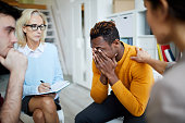 Group of people sitting in circle and sharing their problems and addictions: woman supporting crying black man and touching his shoulder at group psychotherapy session