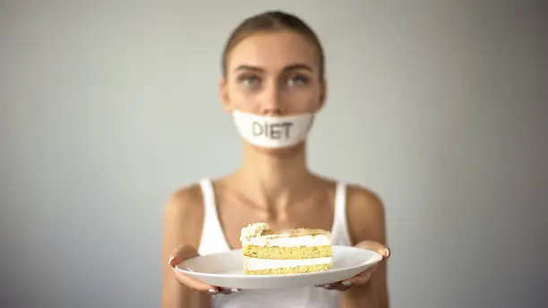 Skinny girl with taped mouth holding cake, refuses to eat sweet, low-carb diet