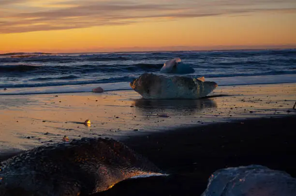 Sunrise on Diamond Beach in Iceland.  Various size icebergs reflecting the morning light, on black sandy beach with ocean waves rolling in.