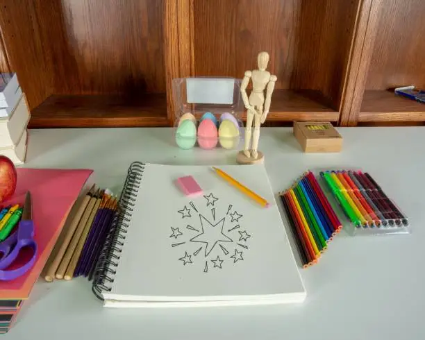 Desk loaded with different drawing materials; markets, crayons, paintbrushes, color pencils, pencil, chalk, and wooden model.