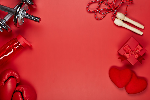 Dumbbells, boxing gloves, rope, bottle for water, gift box and two red hearts on a red background.Top view with copy space. Valentine's Day card. Fitness, sport and healthy lifestyle concept.