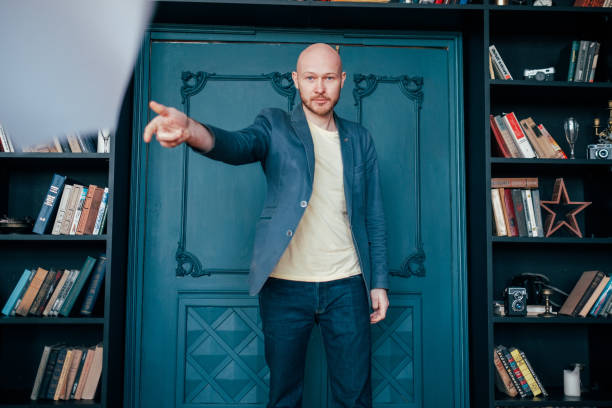 istock Attractive adult successful bald bearded man in suit throwing documents at camera on library book wall blue background 1124331418