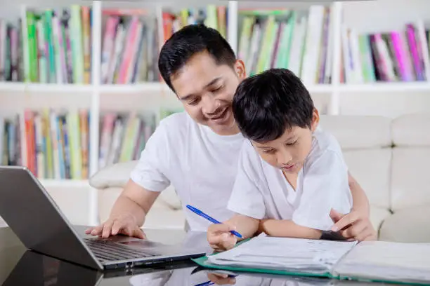 Picture of young father using a laptop while helping his son to study in the library